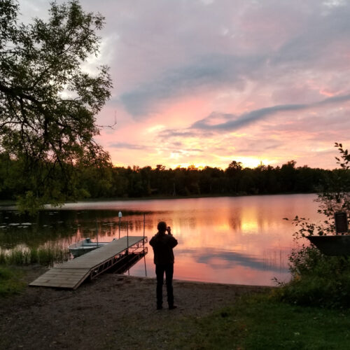 The silhouette of a man standing at the edge of a lake as the sun sets.