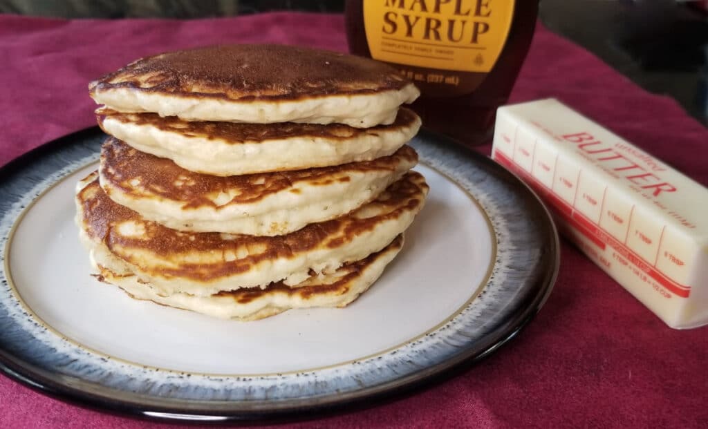 Stacked pancakes on a plate with syrup and butter in the background