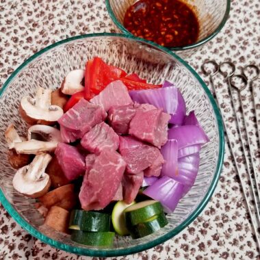 Beef and vegetables in a bowl next to skewers and a kabob marinade.