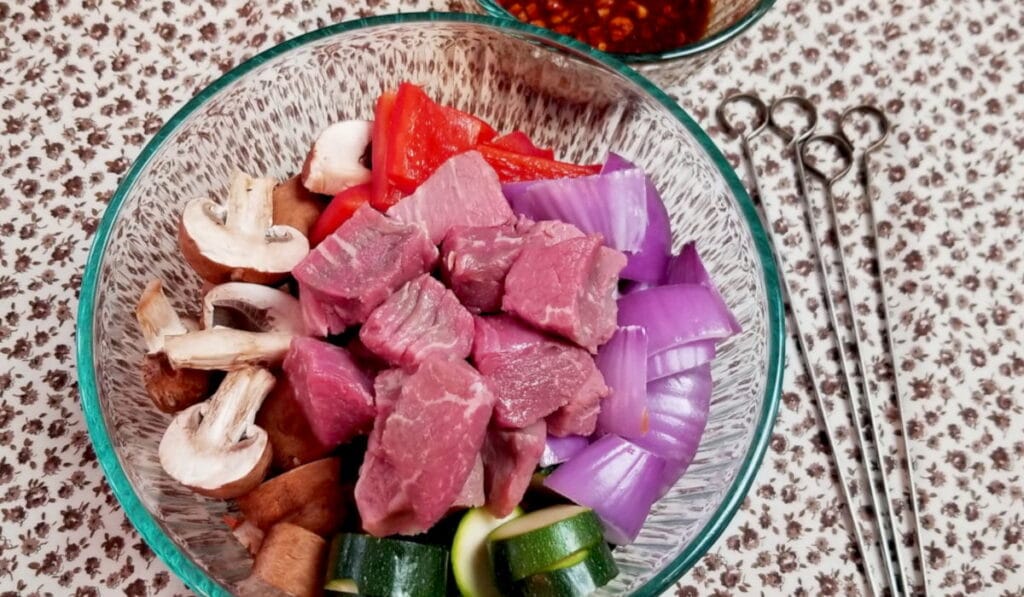 Kabob skewers next to a bowl of  meat and vegetables and another of sauce.