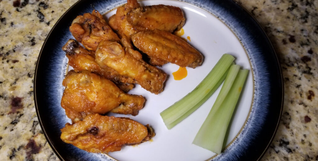 Crispy buffalo wings lay on a plate, dressed with buffalo wing sauce that has lightly spread to the dish. Two pieces of celery fill the empty space. 