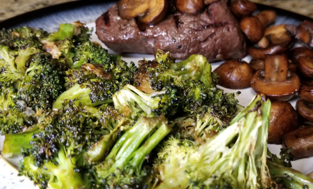 Roasted balsamic broccoli on a plate with a juicy steak and plump mushrooms in the background. 