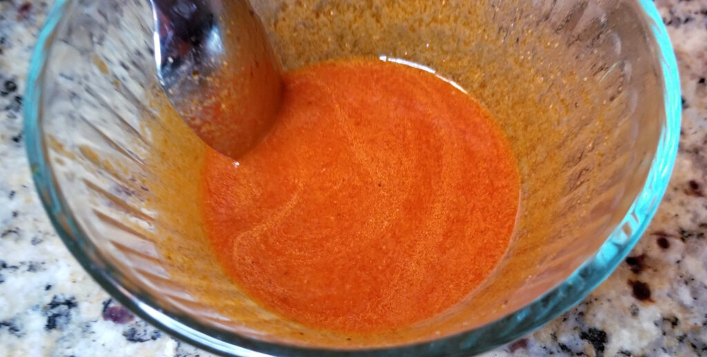 Buffalo wing sauce, in a clear pyrex bowl. A spoon is dipped in the bowl and swirls of butter are visible.
