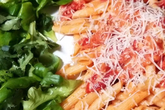 A close up of sun dried tomato pasta next to a mixed greens salad
