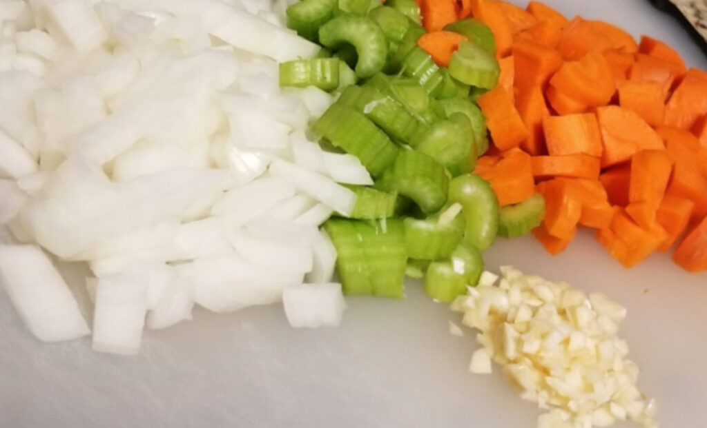 Carrots, celery, onion, and garlic cut up laying on a white cutting board. 