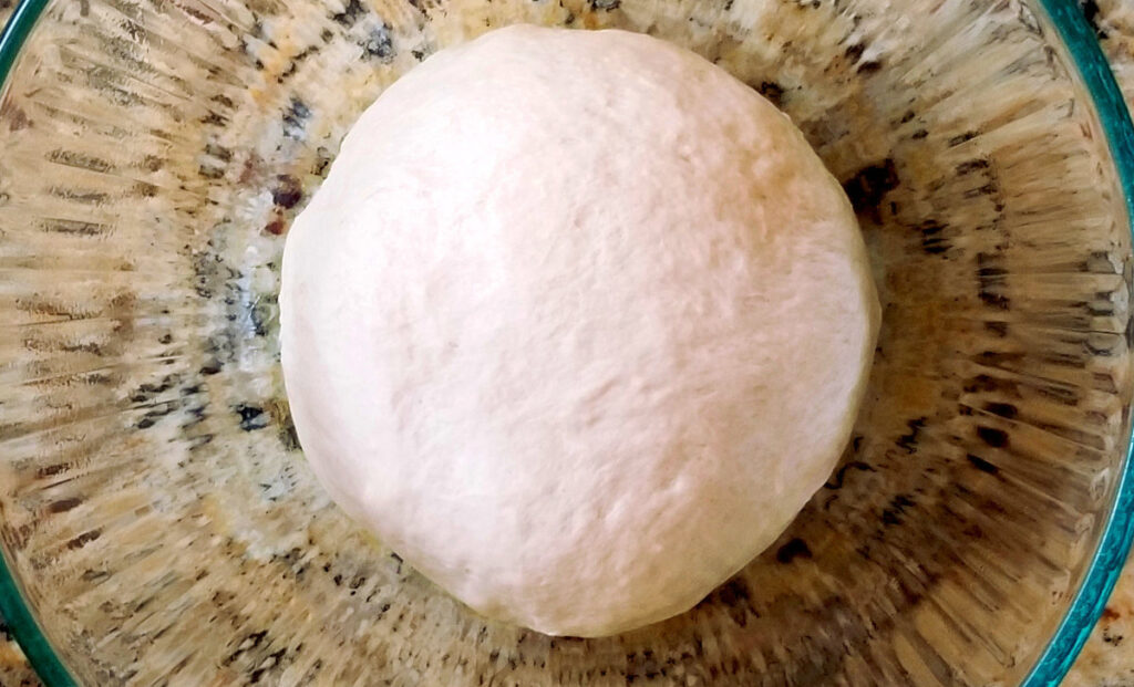 The ball of French loaf dough after its first rise, sitting in a  glass bowl. 