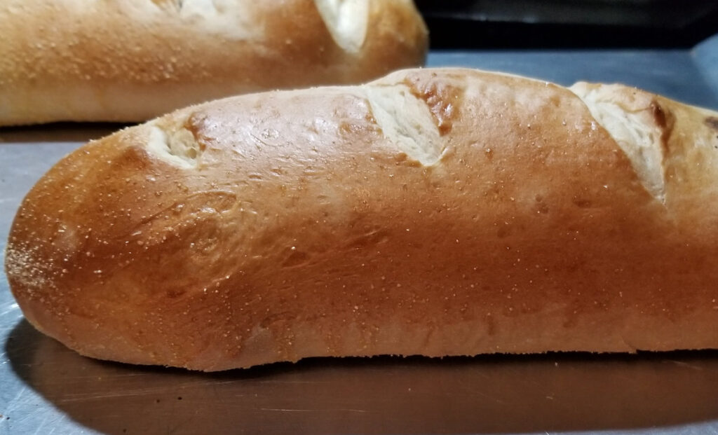 A close up of a French loaf with another in the background, sitting on a baking sheet. 