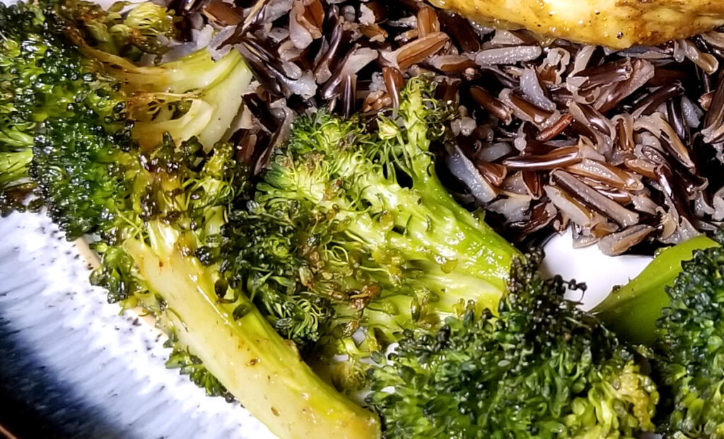 Close up of roasted broccoli on a plate with wild rice with a slight view of the honey curry chicken. 