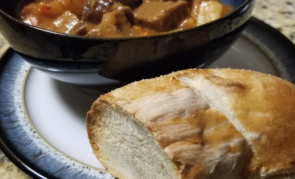 crusty French bread sitting on a plate in front of a bowl of beef stew. 