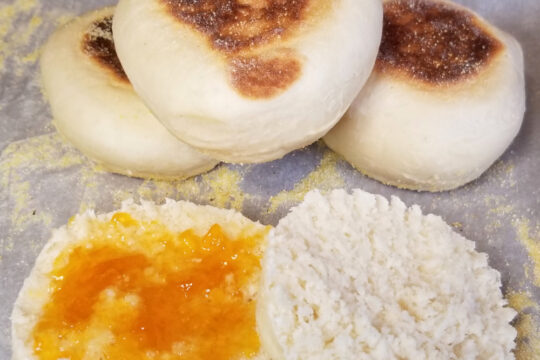 stacked english muffins in the background and an open muffin in the front with apricot jam