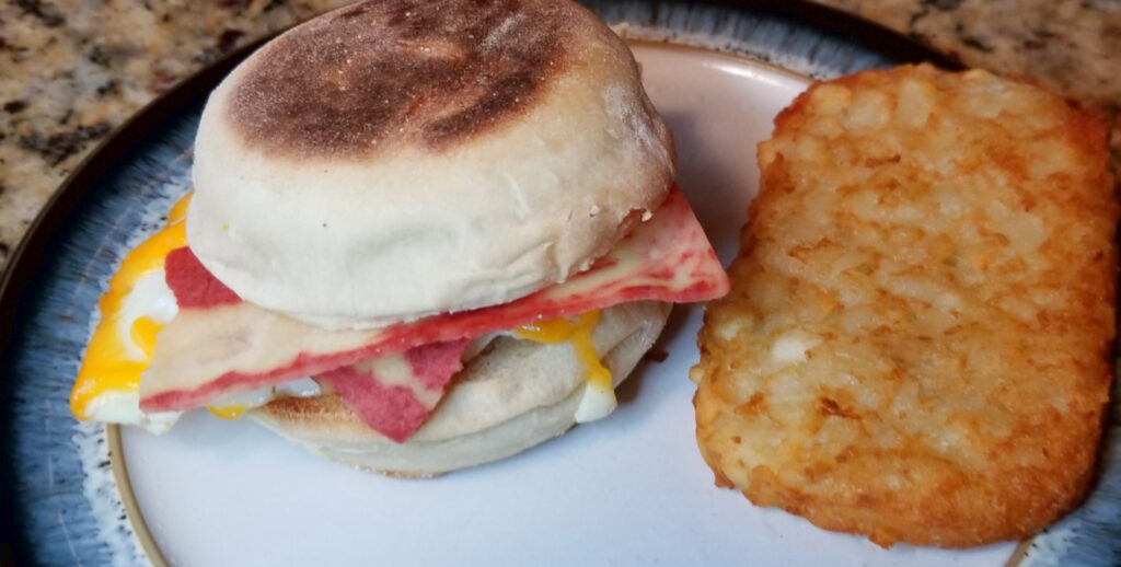 An English muffin sandwich with fake bacon and cheese next to a hashbrown patty on a blue and white plate.