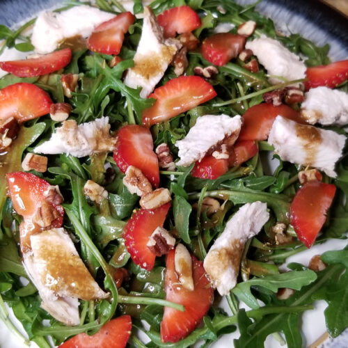 A close up of strawberry, chicken, and arugula salad with a balsamic dressing drizzle