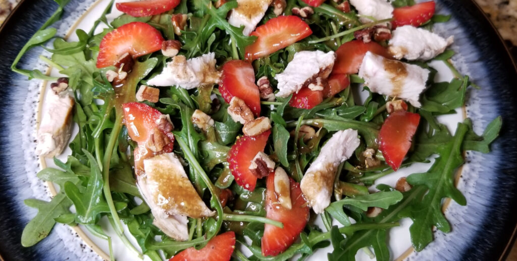 Arugula, strawberries, chicken, and pecans on a plate and drizzled with balsamic dressing.
