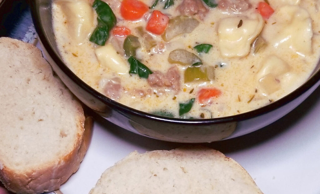 A bowl of sausage and spinach tortellni soup on a plate with two slices of french bread. 