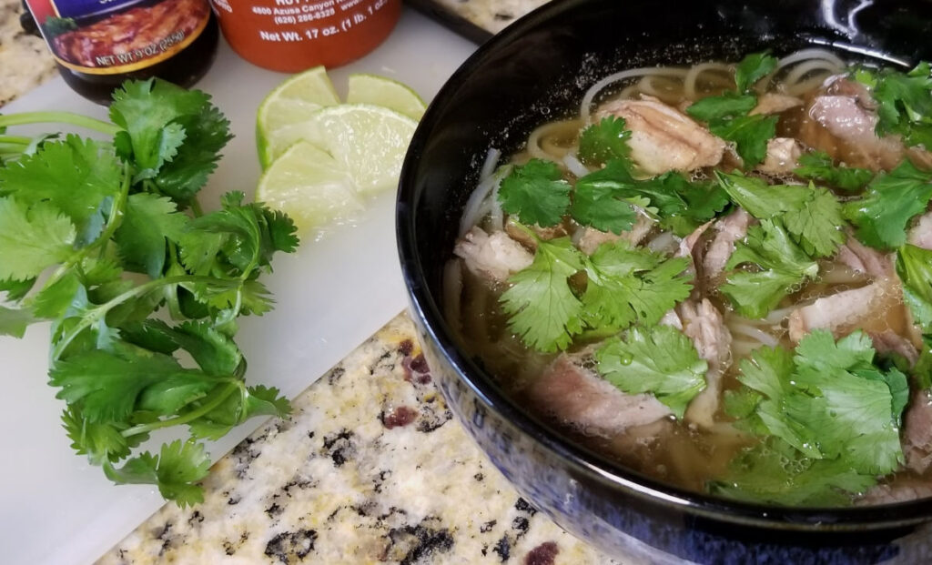 a bowl of pho next to a cutting board with cilantro and cut lime