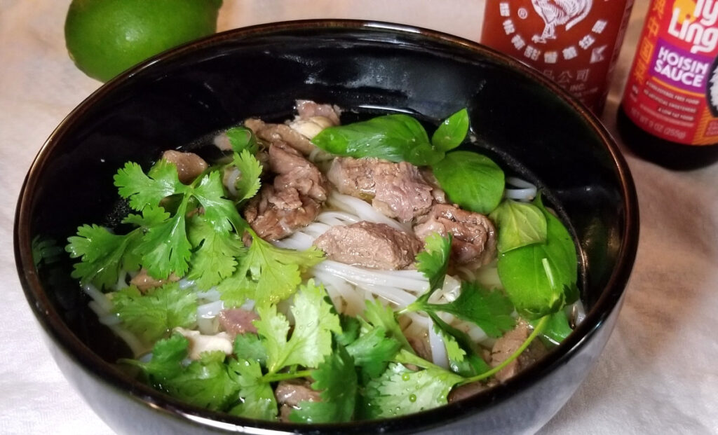 Pho topped with herbs, in a large bowl, with sriracha, hoisin sauce, and lime in the background.