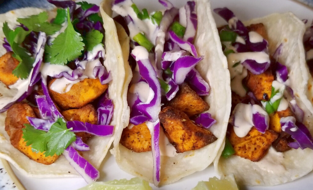Close up of sweet potato tacos with red cabbage, cilantro, and green onion. A small bit of lime is visible in front of the tacos.