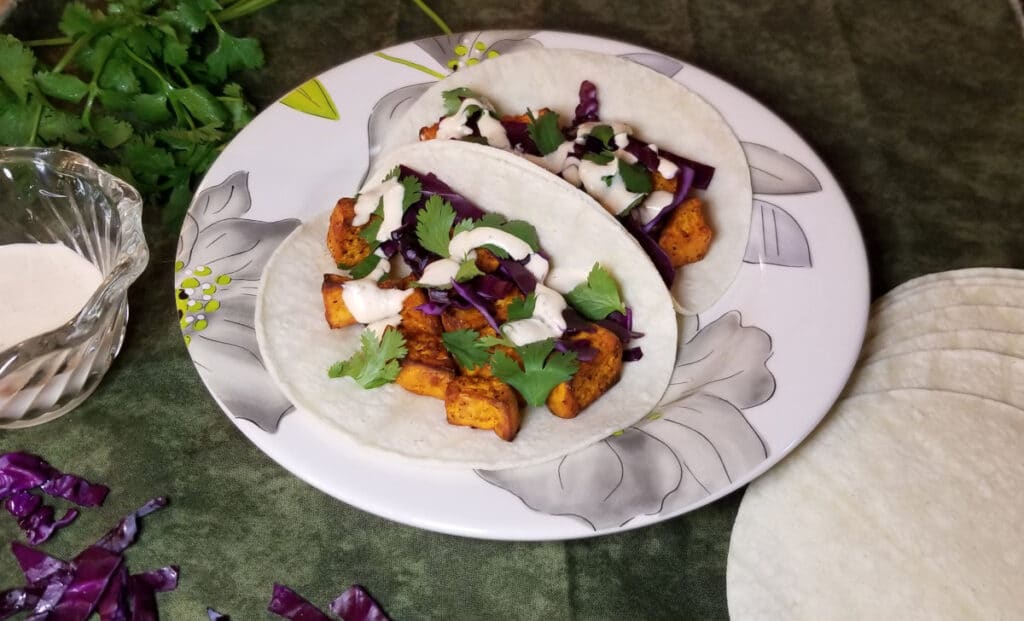 Sweet potato tacos on a plate with tortillas, creamy sriracha sauce, cabbage, and cilantro in the background