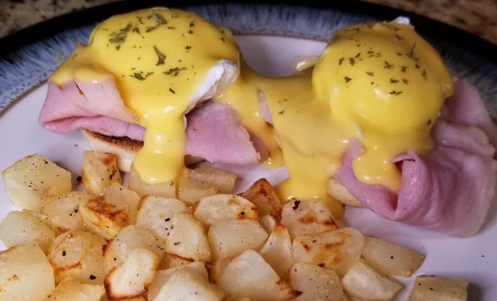 Poached eggs over ham with hollandaise and sprinkled herbs, next to crispy potatoes. 
