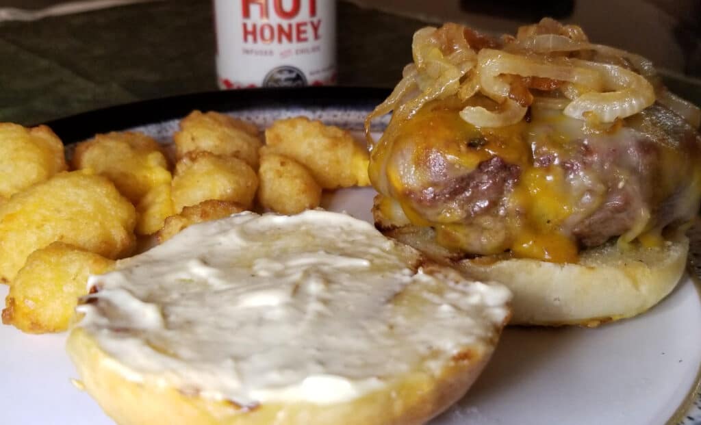 A juicy caramelized onion cheese burger on a plate with cheese curds and hot honey in the background. 