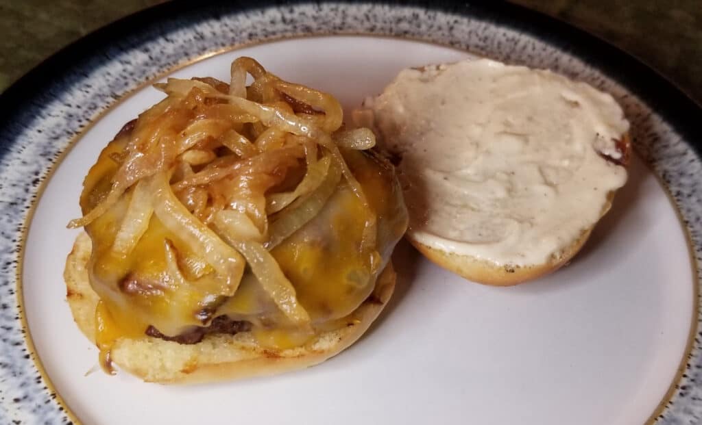 An open-faced hamburger with cheese and caramelized onions on a plate, with aioli on the top bun. 