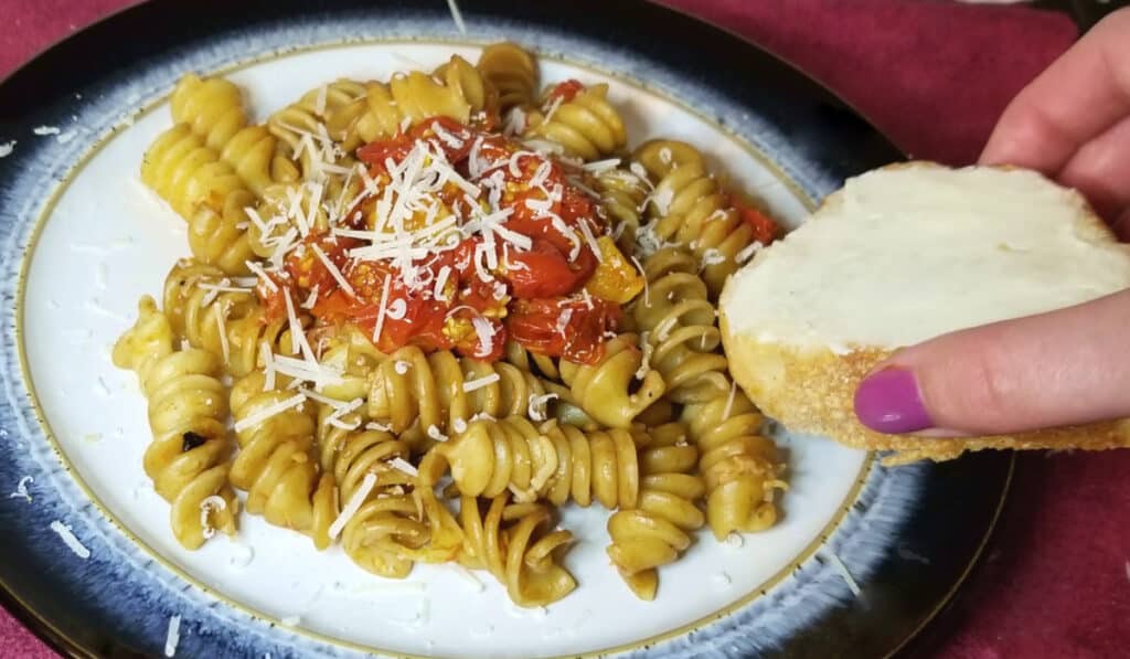 Burst tomato sauce over rotini pasta being dipped into with a slice of buttered French bread. 
