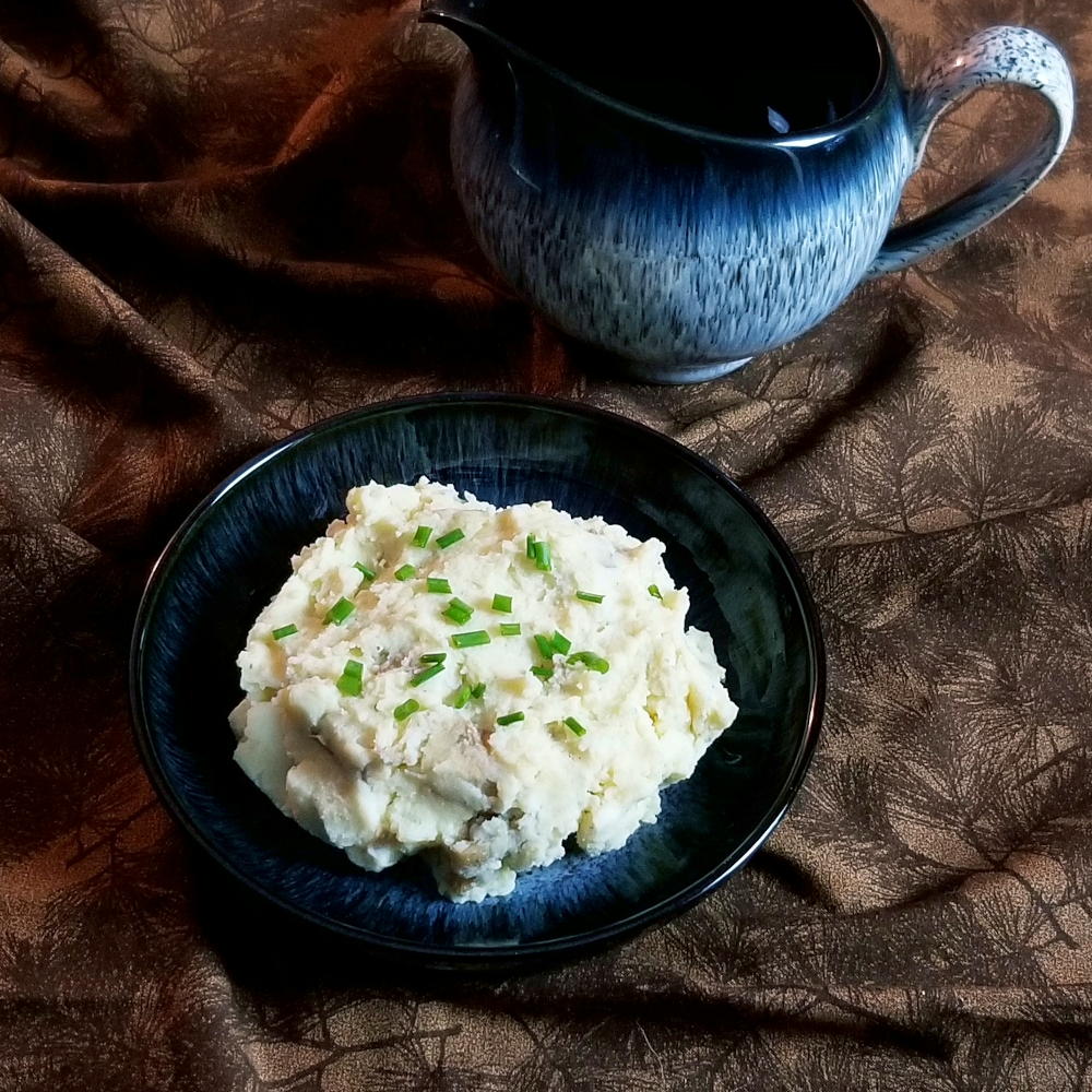 Mashed sour cream and chive potatoes in a bowl with a gravy boat in the background.