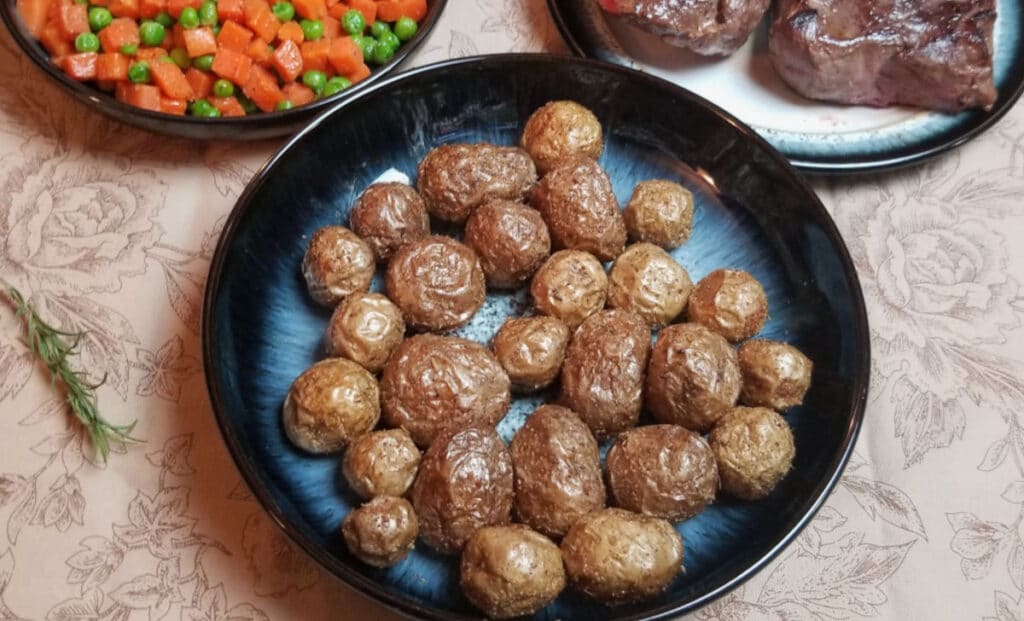 A bowl of air fried baby potatoes with rosemary, peas and carrots, and steak in the background. 